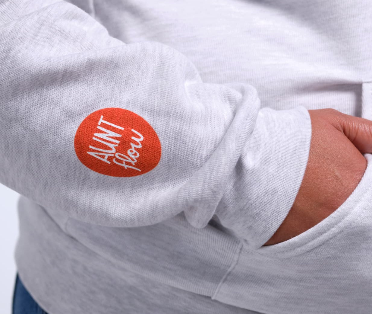 Close-up of a person wearing a light heather gray hoodie, showing a red circular sleeve patch with the white text 'AUNT Flow'.