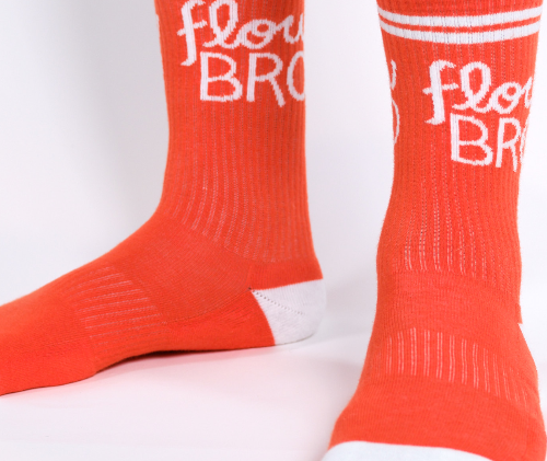 A pair of orange cotton crew socks with the text 'FLOW BRO' written across the top, featuring a classic ribbed design and placed on a person's feet.